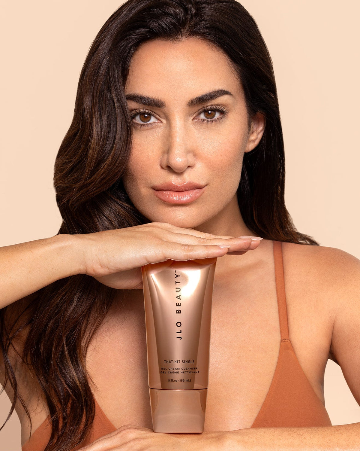 A woman with long dark brown hair holds a tube of JLO Beauty Clear Gel Cleanser in front of her face against a solid beige background.