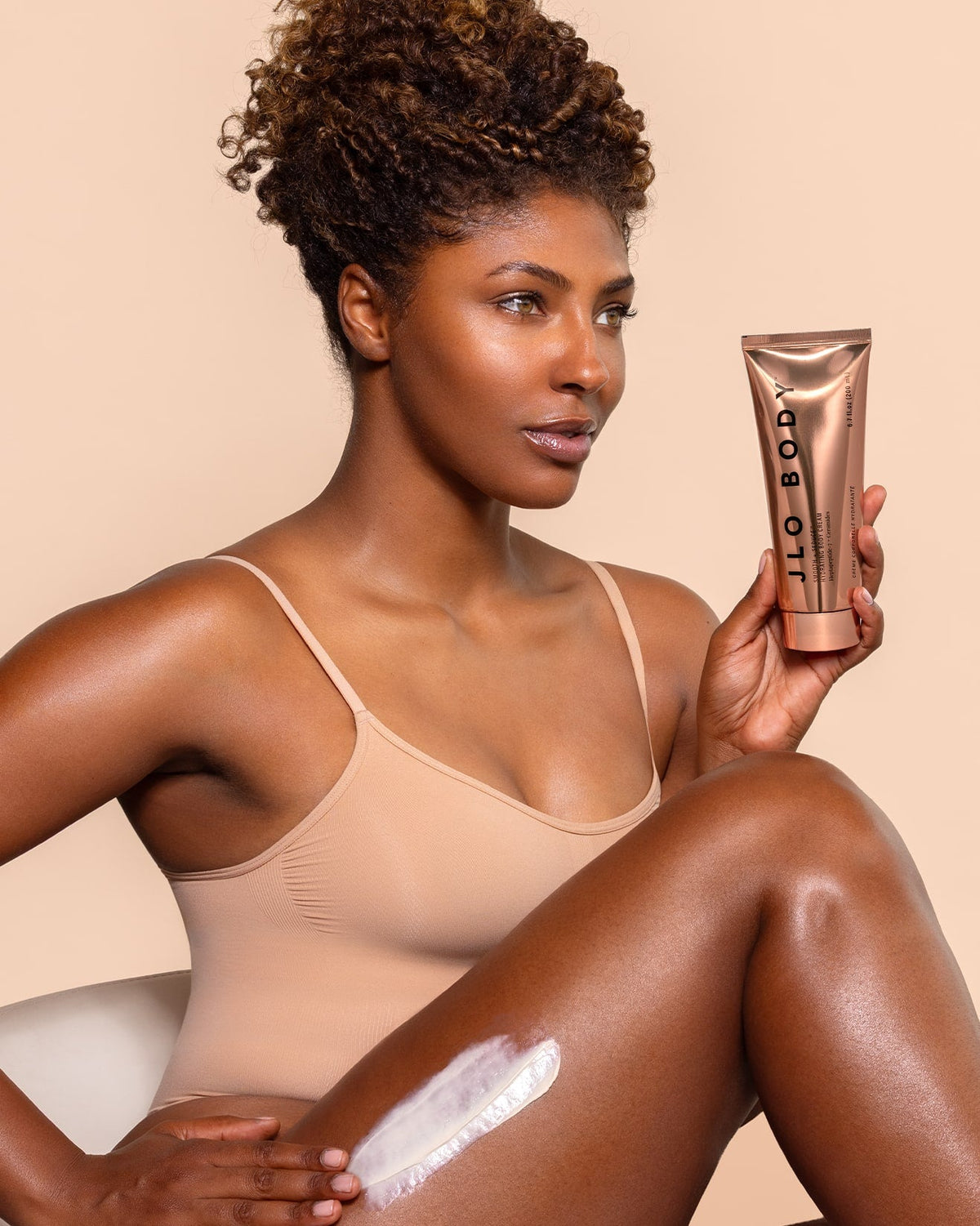 A woman holding a metallic rose gold tube of 'JLO body' lotion that she is applying to her leg